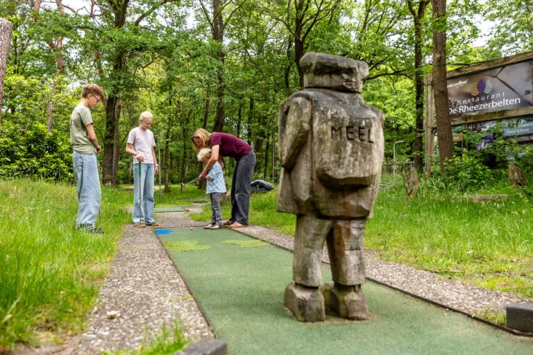 Gezin speelt midgetgolf bij De Rheezerbelten met houten sculptuur op de voorgrond en restaurantbord op de achtergrond